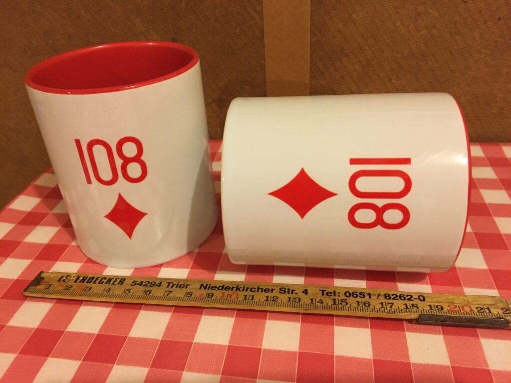 Zwei weiße Tassen mit rotem Aufdruck "Karo 108" auf dem Küchentisch des Autors. Ein steht, eine liegt und ein Zollstock zeigt die Größe.