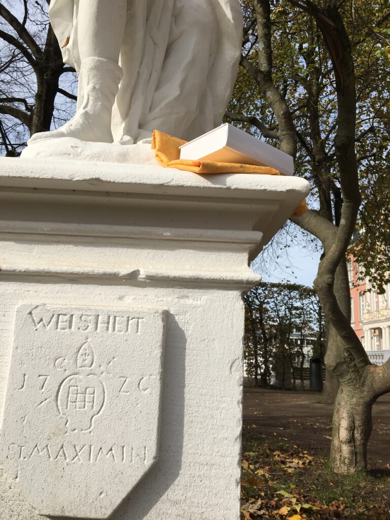 Bild der Weisheits-Statue beim Kurfürstlichen Palais Trier mit einem Band der "Zehn keinen Bücherlein".